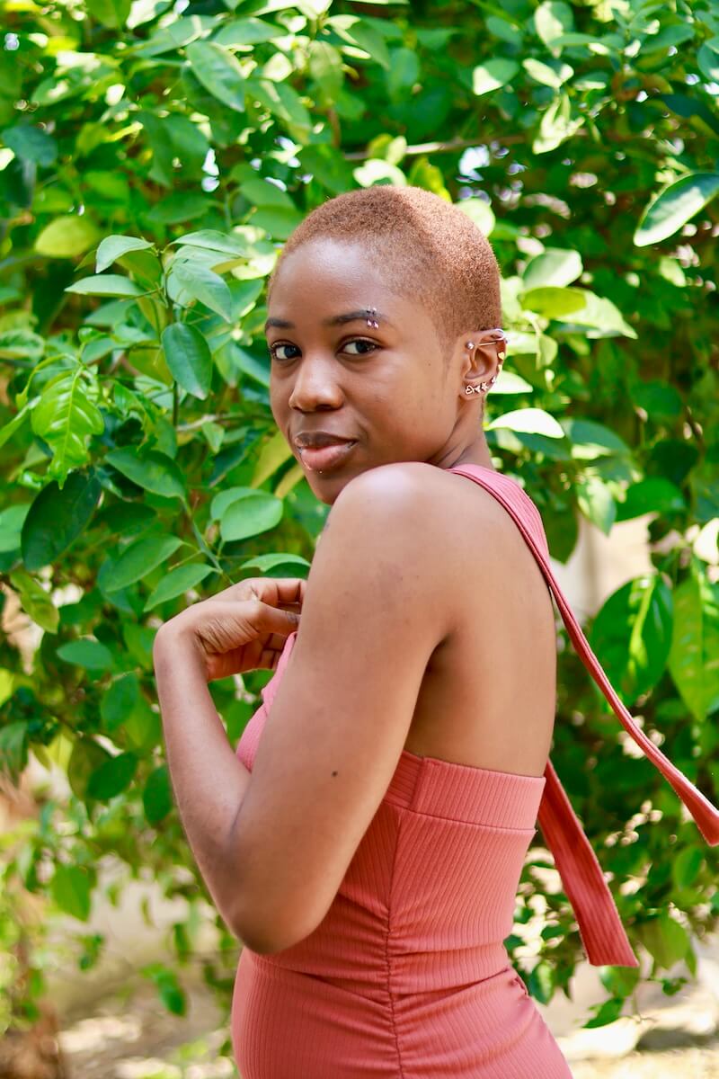 a portrait photo of a woman in a burnt orange dress, standing sideways in front of a tree with lush green leaves behind her, facing the camera from said side